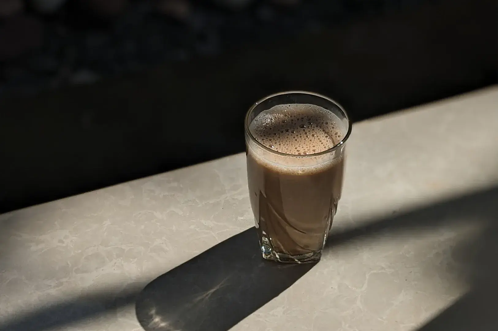 Clear glass tumbler filled with frothy milk tea or chai resting on a sunlit marble ledge.