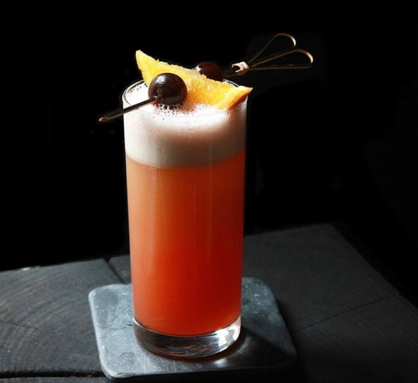 an orange cocktail in a hi-ball glass garnished with fruit on a skewer