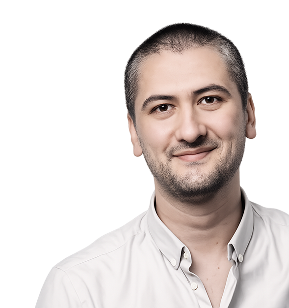 A man with short dark hair and light stubble smiles warmly against a stark black background, wearing a white button-up shirt — the high-contrast studio lighting giving the portrait a professional, confident tone.