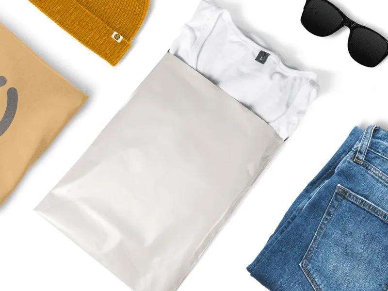 a gray shipping bag placed at the center, partially open with a white t-shirt peeking out. Surrounding the bag are various clothing accessories, including a mustard-yellow knitted beanie, a pair of black sunglasses, a folded pair of blue denim jeans, and a beige tote bag with a subtle design.