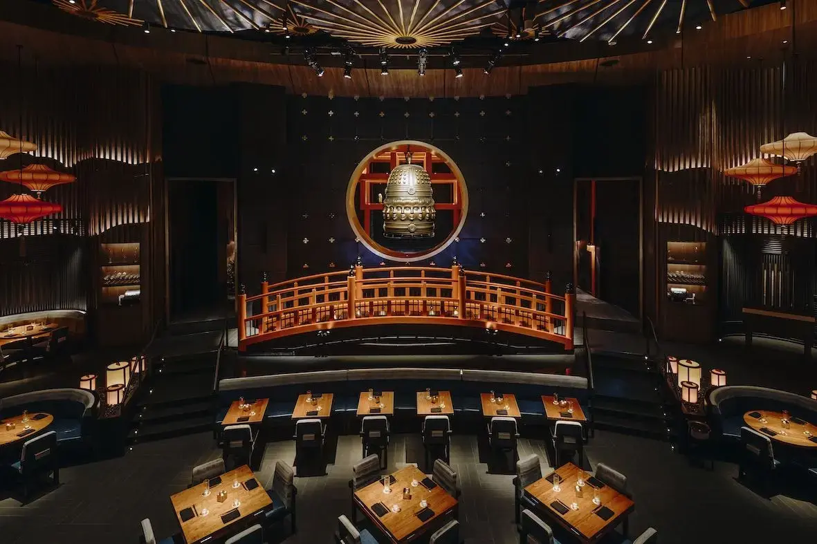 This image showcases a dramatic, high-ceilinged dining room featuring a prominent orange footbridge and a large bronze bell suspended within a circular backlit frame. The atmosphere is upscale and moody, accented by warm lantern lighting, wood-paneled walls, and neatly arranged tables with minimalist place settings.