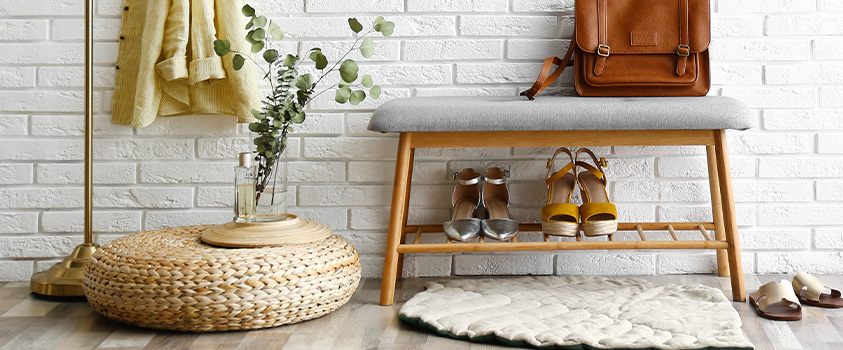 An Artiss Bamboo Shoe Storage Bench holding two pairs of shoes and a leather handbag. On the left is a straw pouf holding a vase of greenery and a perfume bottle, and a brass coat rack. It is in an entryway with a light wood floor and white brick wall. 