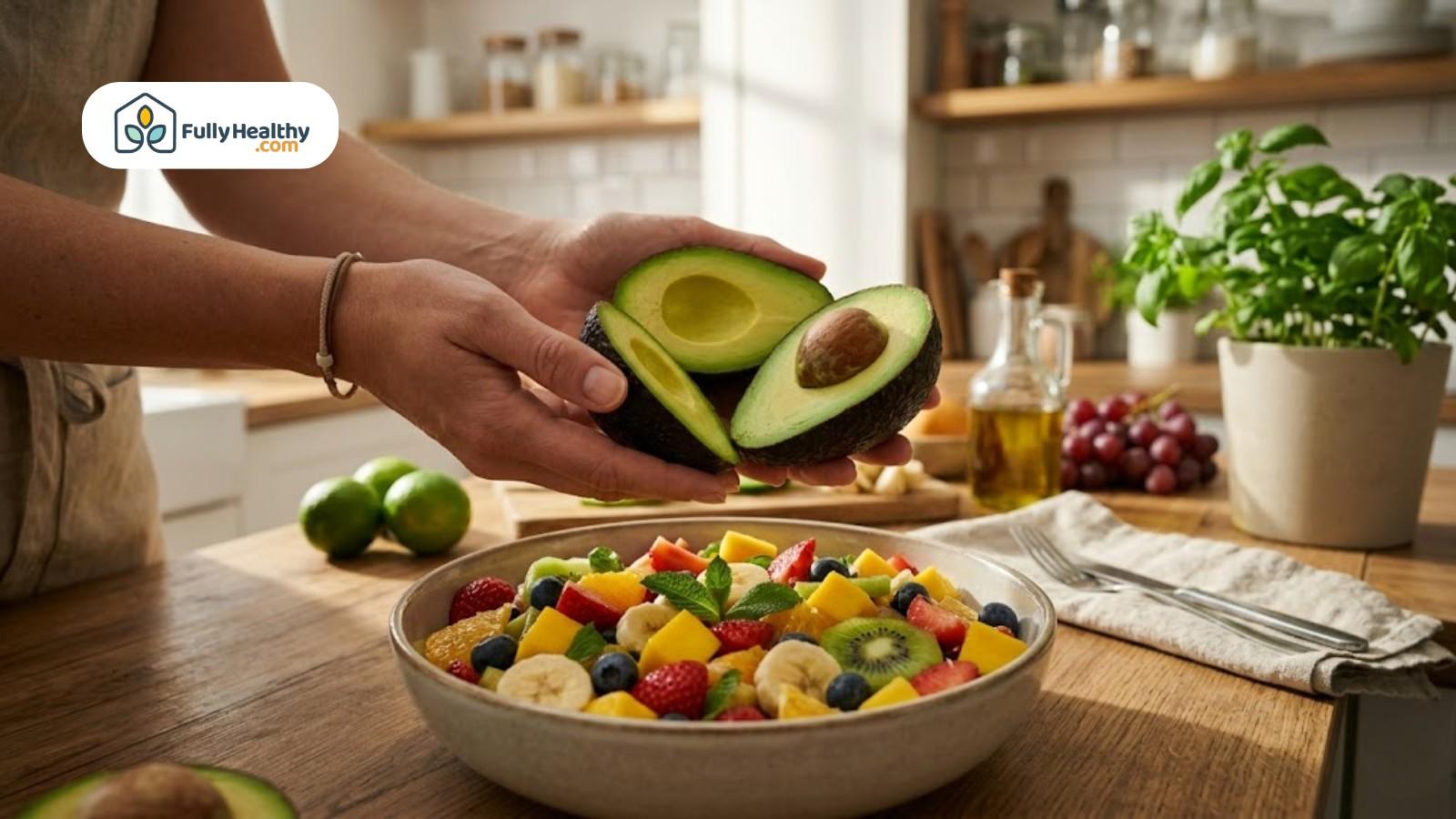 Hands holding avocado halves above colorful fruit salad in kitchen setting
