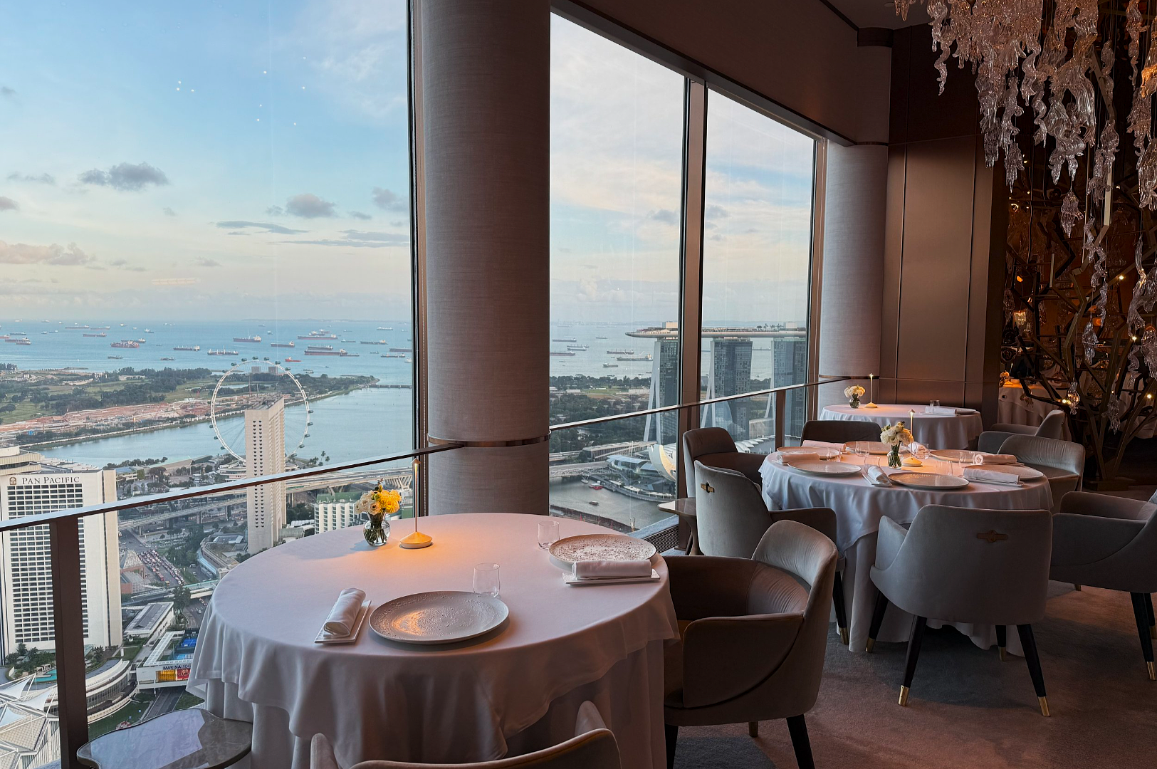 Luxurious restaurant with elegantly set tables by large windows, offering a panoramic city and sea view. The atmosphere is serene and sophisticated.