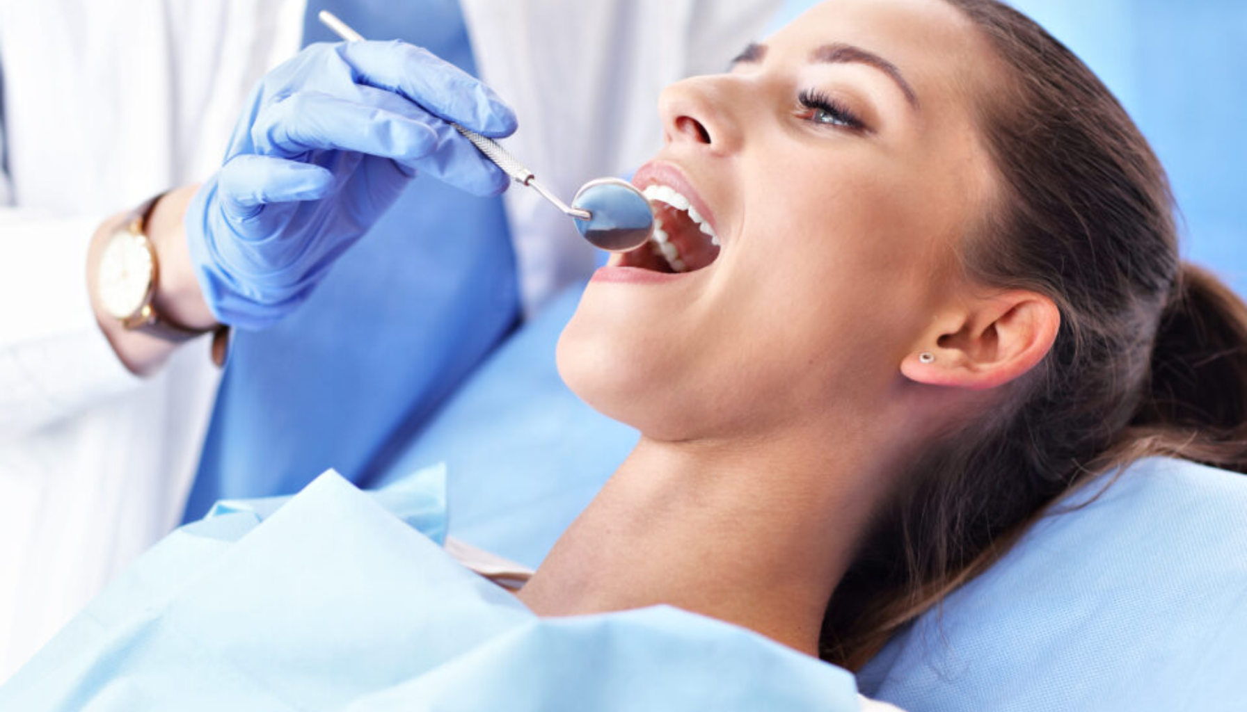 Kvinne ligger i tannlegestol mens tannlege undersøker munnen med speil og hansker.