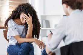 A woman with curly hair sits with her head in her hand, appearing distressed, during a therapy session.