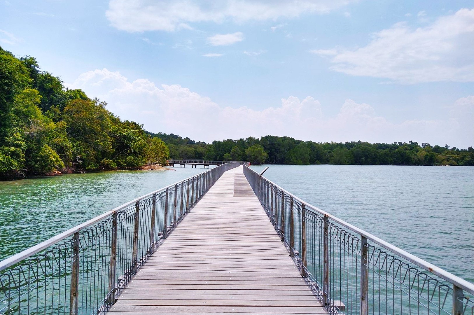 Wooden boardwalk stretches across calm water, leading to lush green trees under a partly cloudy sky, evoking tranquility and serenity.