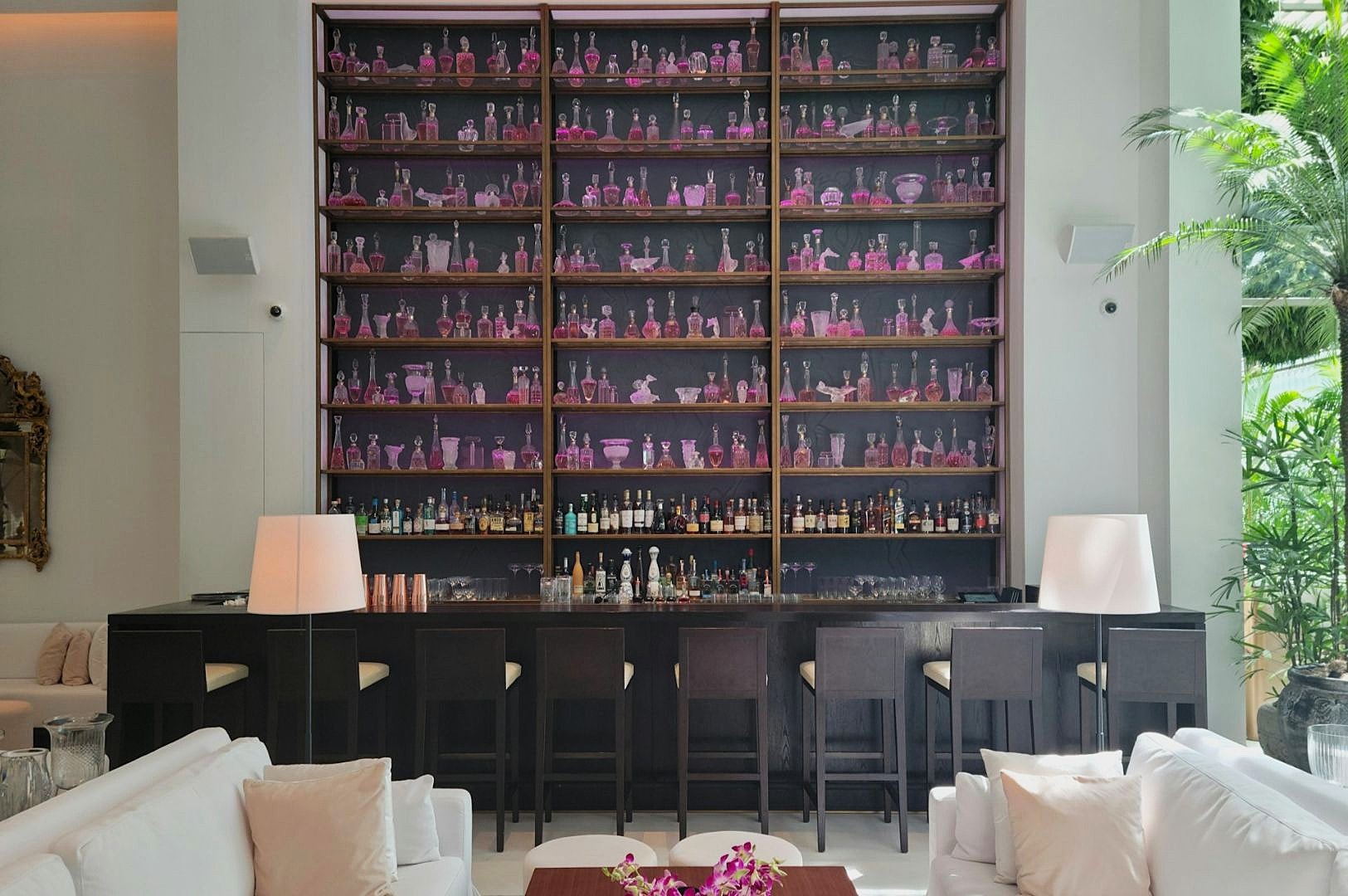 Elegant bar with tall shelves displaying purple glassware. Dark bar stools and seating area create a modern, sophisticated ambiance with soft lighting.