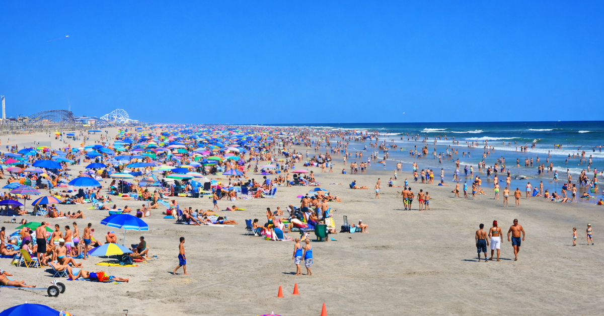 Crowded summer beach in North Wildwood NJ with colorful umbrellas, families enjoying the sand and surf, and boardwalk amusement rides in the distance.