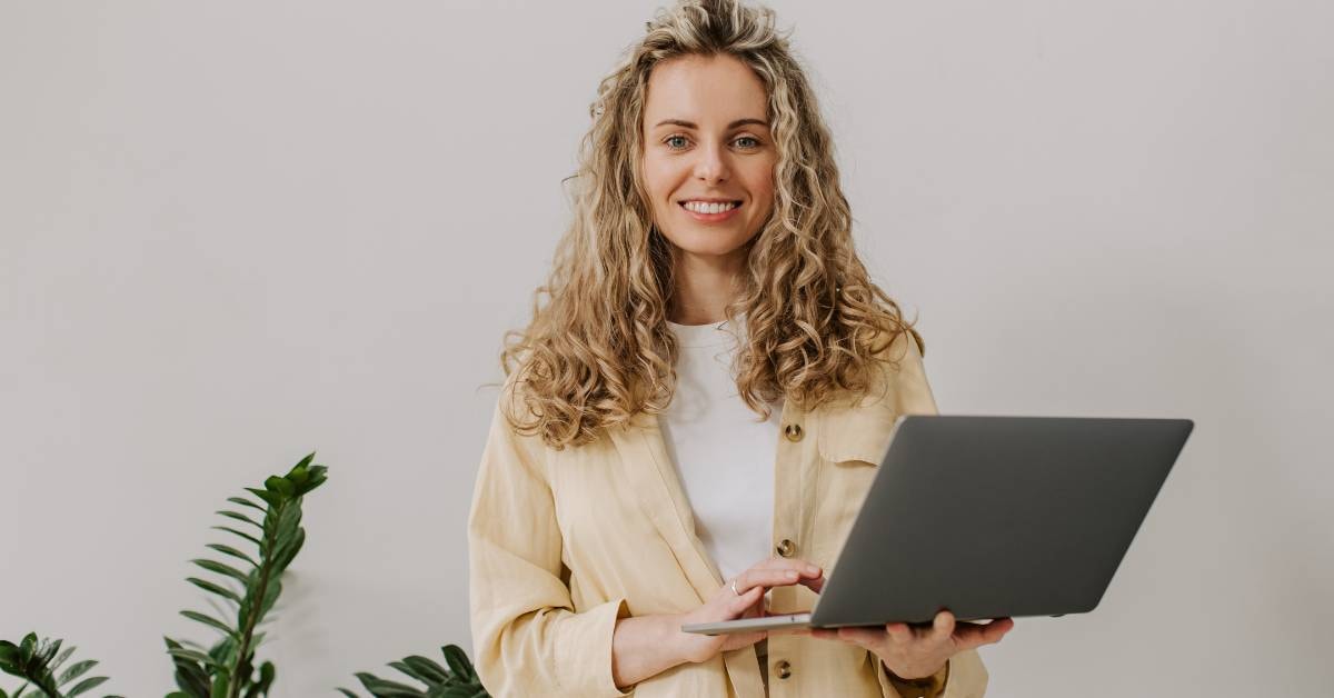 A smiling woman with curly blonde hair holding a laptop, standing in a bright room with plants, preparing to apply for her business tax certificate.