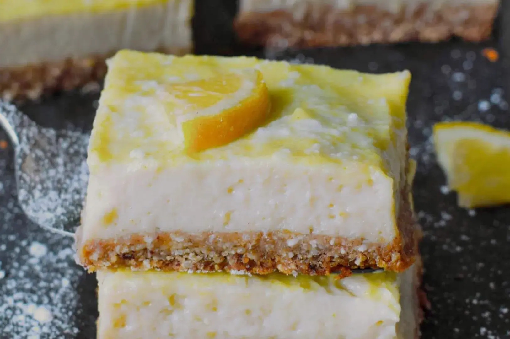 A creamy lemon cheesecake bar with a golden crumb crust and orange slice sits on a dark plate dusted with powdered sugar, inviting and bright.