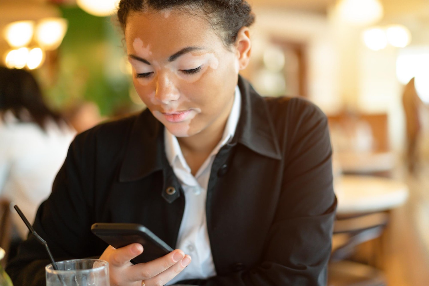 A woman with vitiligo, dressed in business attire, sits at a café table focusing intently on her smartphone. She appears engaged in professional communication, highlighting the role of SMS for business communication and customer communication tools.