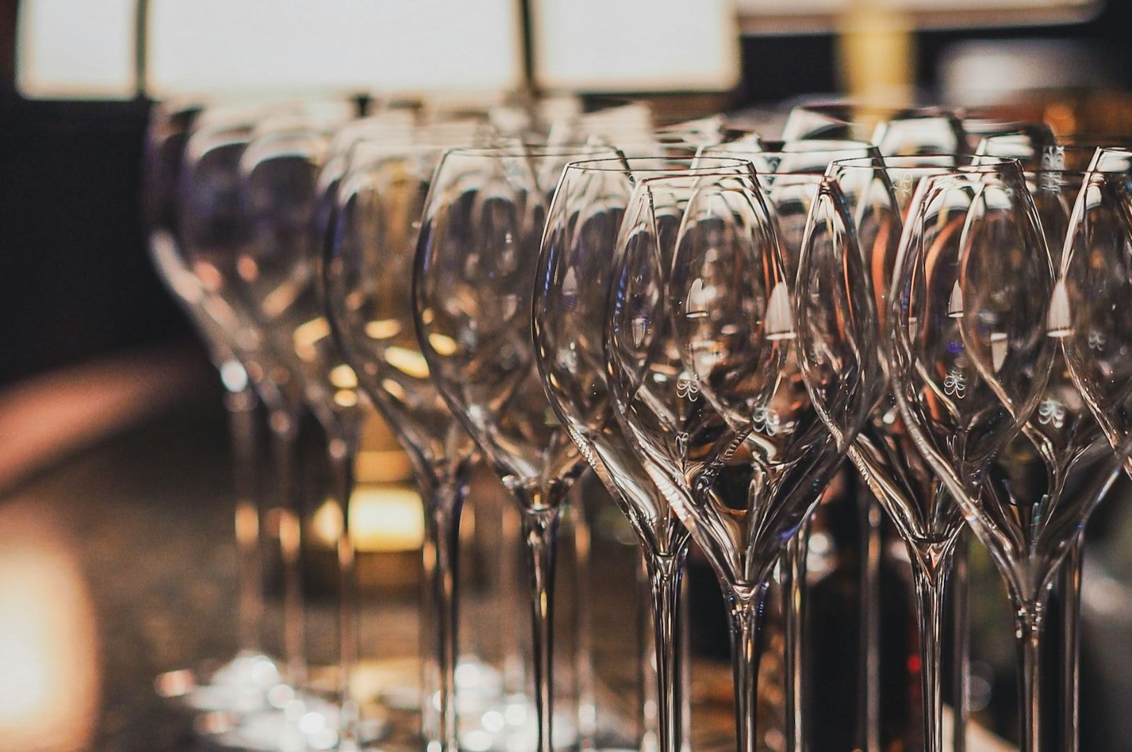 Rows of empty, elegant wine glasses on a polished surface. The dim lighting and reflections create a warm, inviting atmosphere with hints of luxury.