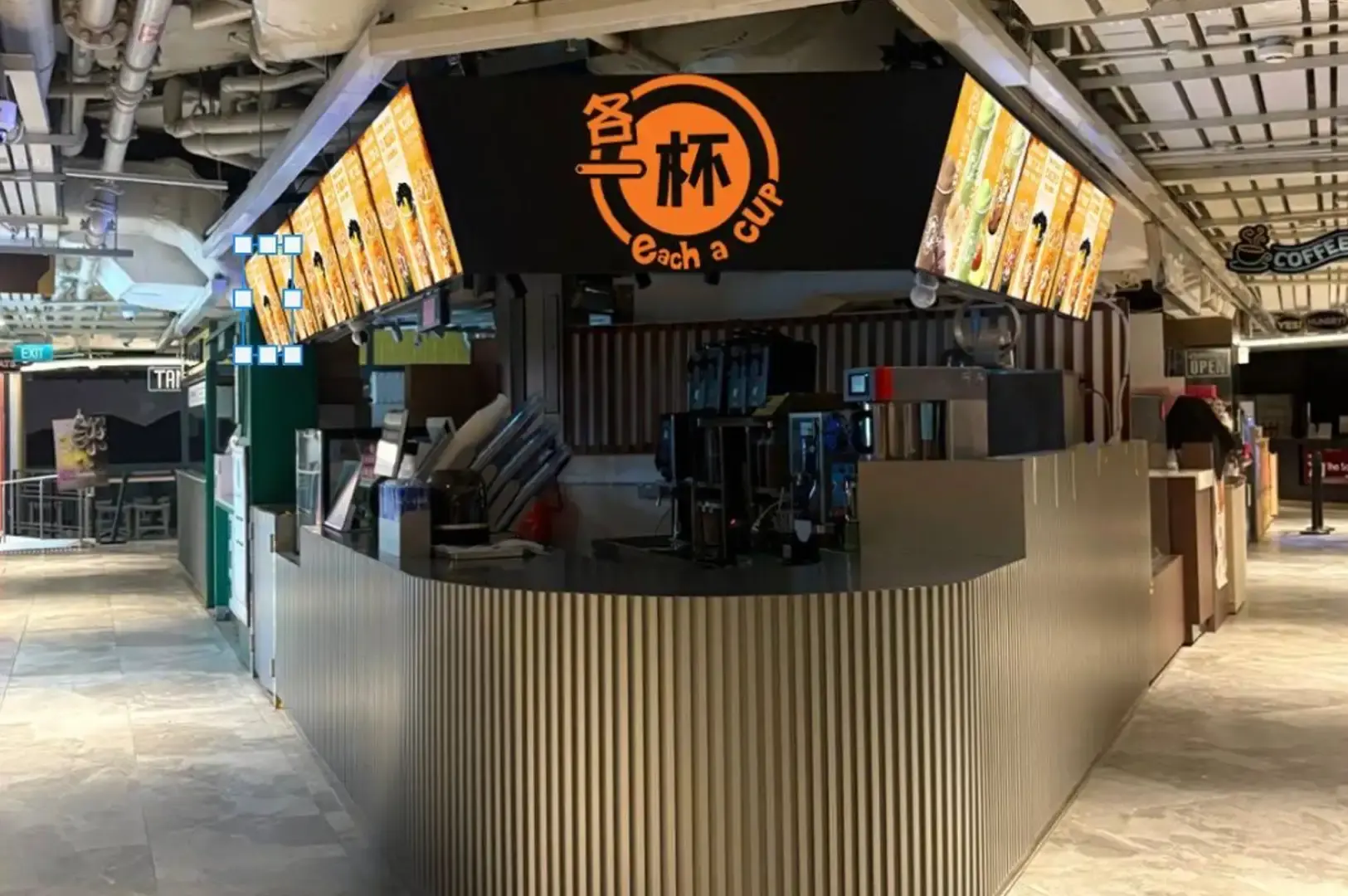A modern, standalone Each A Cup kiosk booth situated in a mall hallway, featuring sleek corrugated metallic paneling along its curved counter base, illuminated overhead digital menu displays, and a prominent black-and-orange brand sign.