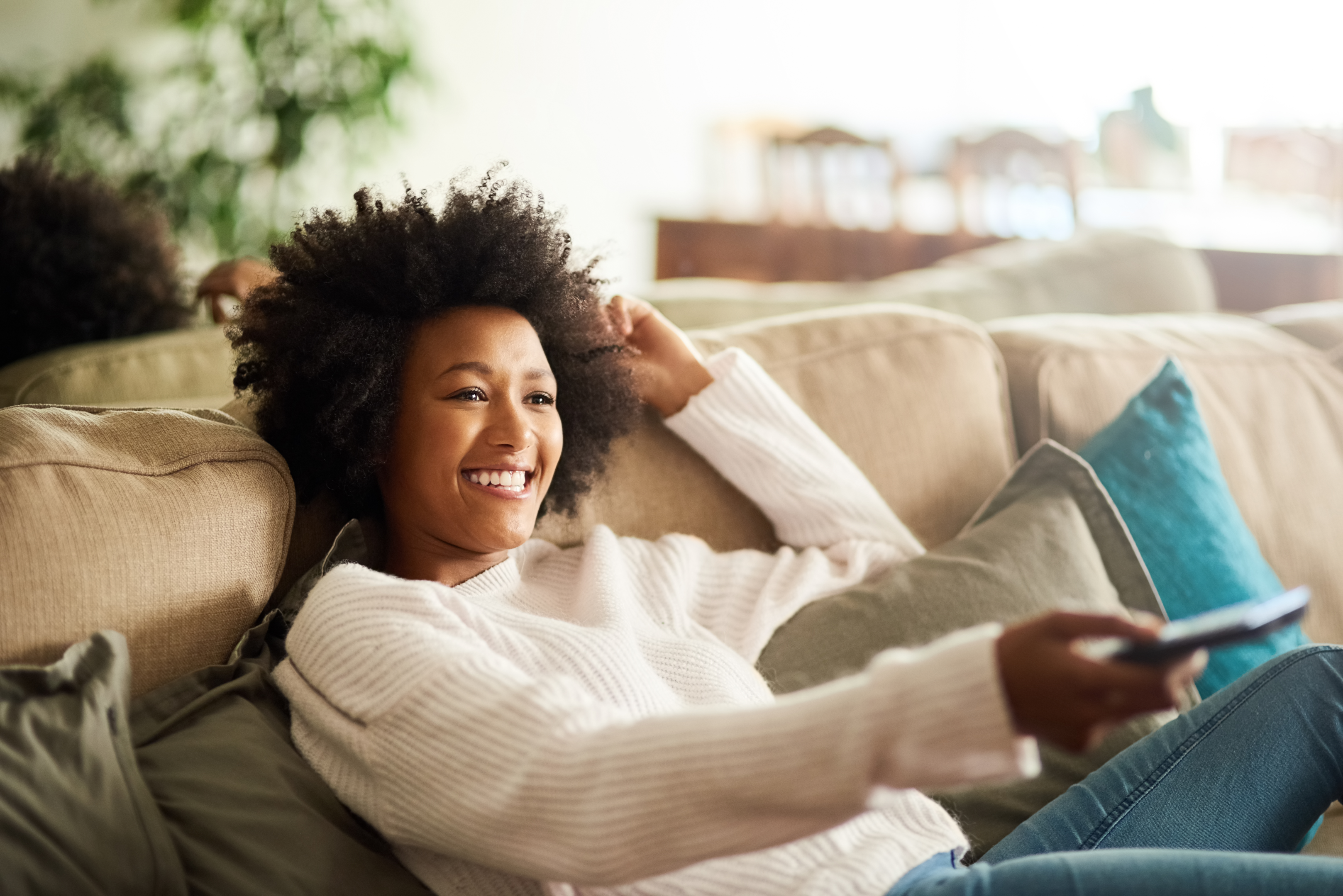a woman smiles on her living room couch while holding a tv remote