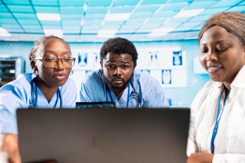 A diverse team of three healthcare workers in scrubs and lab coats intently reviews a laptop screen in a hospital environment. Unified Communications and real-time team collaboration tools drive seamless communication and support business productivity in diagnosis, treatment planning, and patient care coordination.