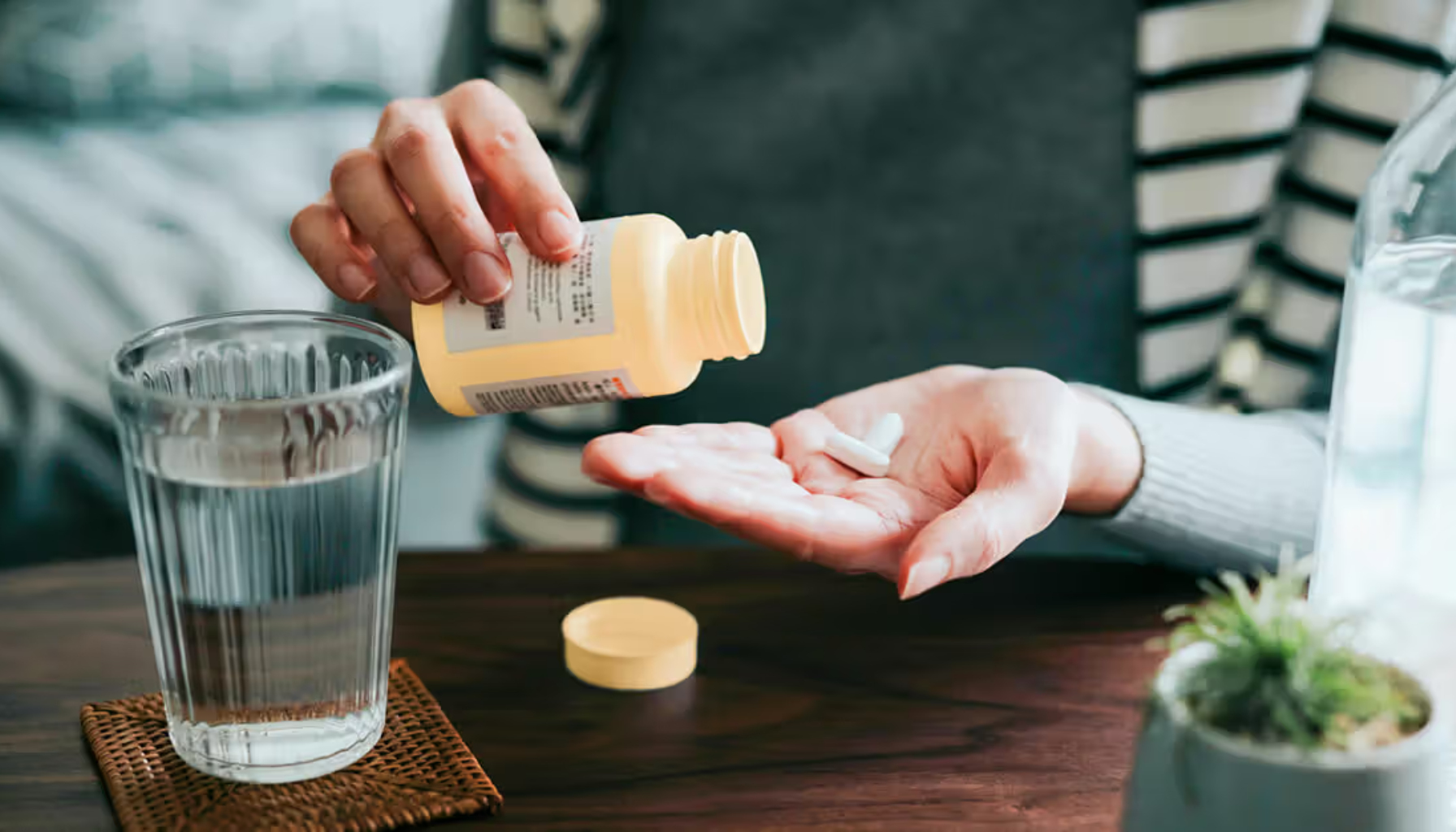 Person heller kosttilskudd i hånden med vannglass på bordet, klar til å ta tabletter eller vitaminer hjemme i en rolig setting.
