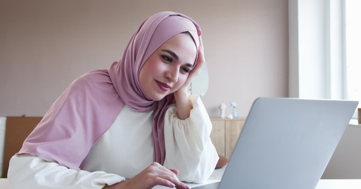 A woman in a pink hijab thoughtfully working on a laptop, researching what happens if you don't file taxes for OnlyFans.