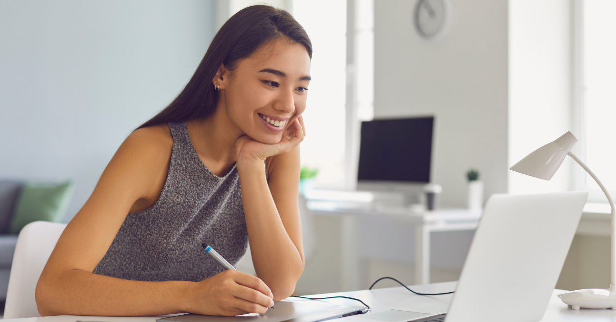 A smiling woman working on a laptop in a bright office; exploring differences between audit vs tax roles.