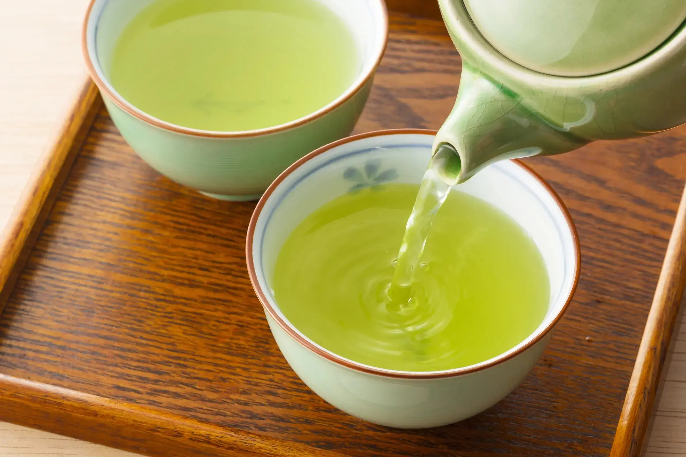 A light green tea is poured from a matching ceramic teapot into a delicate cup, creating small ripples on the surface of the liquid. The scene is set on a warm wooden tray, featuring a second filled cup and a soft, minimalist aesthetic.
