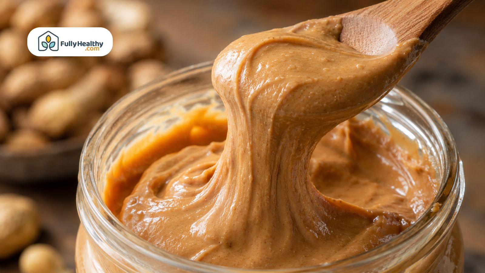 Wooden spoon lifting creamy natural peanut butter from a glass jar close-up.