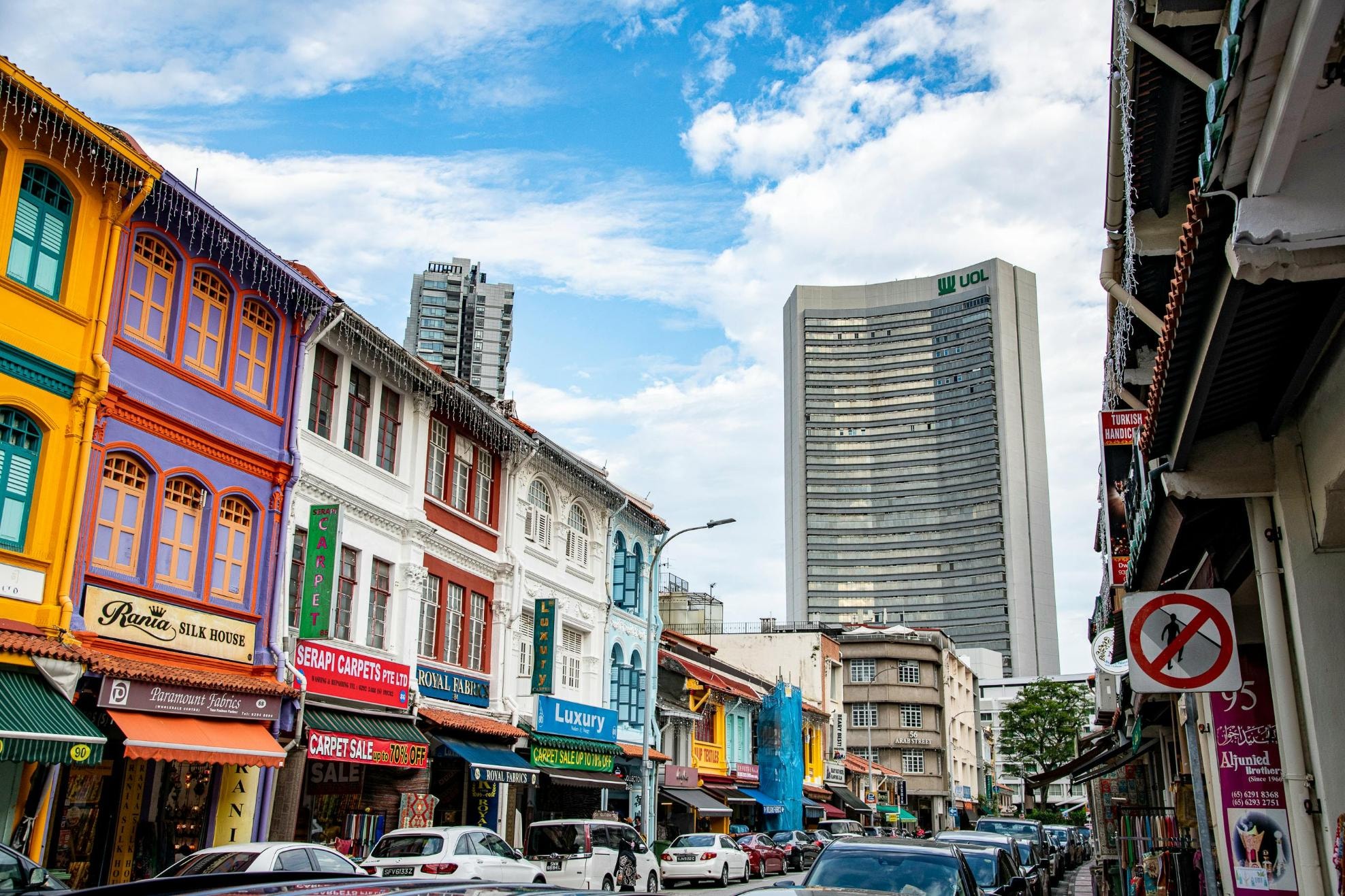 A vibrant row of multi-colored historic shophouses, featuring businesses like silk and carpet stores, lines the left side of a busy city street. Behind these traditional structures, towering modern skyscrapers, including the prominent UOL building, rise against a bright, cloudy blue sky.