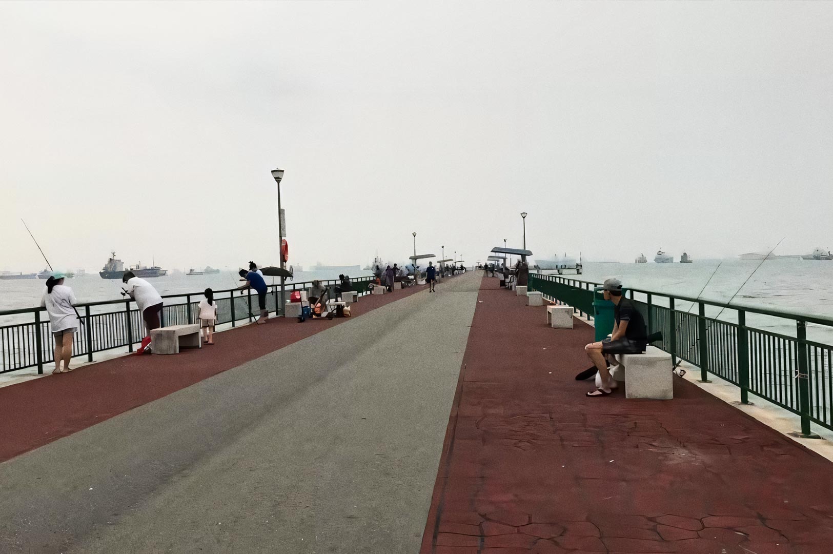 Long pier at Bedok Jetty extending into the sea, with people fishing along both sides. The sky is overcast, and ships are visible in the distance across the water.