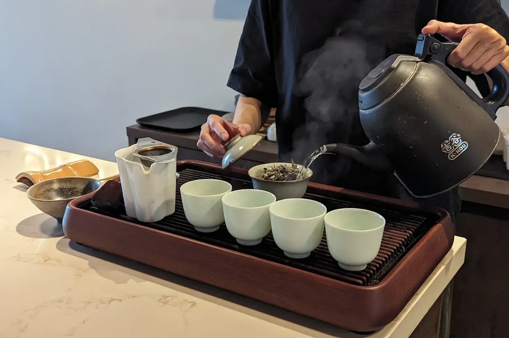 Tea master pouring steaming water from a black kettle into a ceramic gaiwan during a tea service.