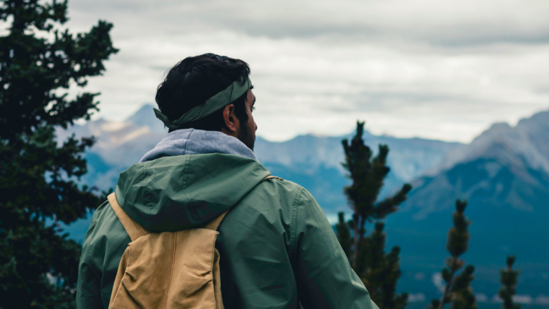 A person in a green jacket and a tan backpack gazes at the mountains under clouds.