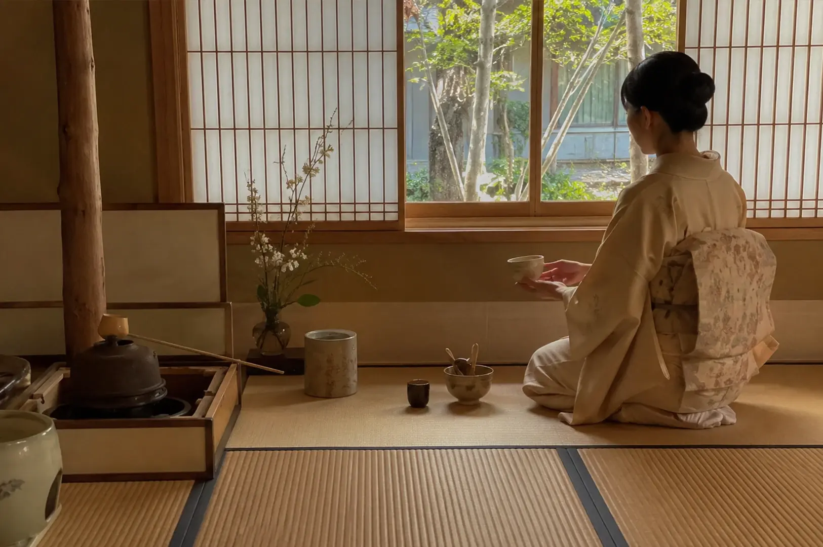 Tea Ceremony Kyoto A Slow Dance of Bowls and Breath.webp," a woman in a kimono is seen from behind, kneeling on tatami mats as she prepares for a tea ceremony. She holds a small tea bowl, looking out through large, grid-paned sliding windows toward a tranquil, sunlit garden. The scene is quiet and minimalist, featuring a simple tea setup on the floor, a vase with delicate flowering branches, and a rustic iron kettle to her left, creating a moment of stillness and contemplation.