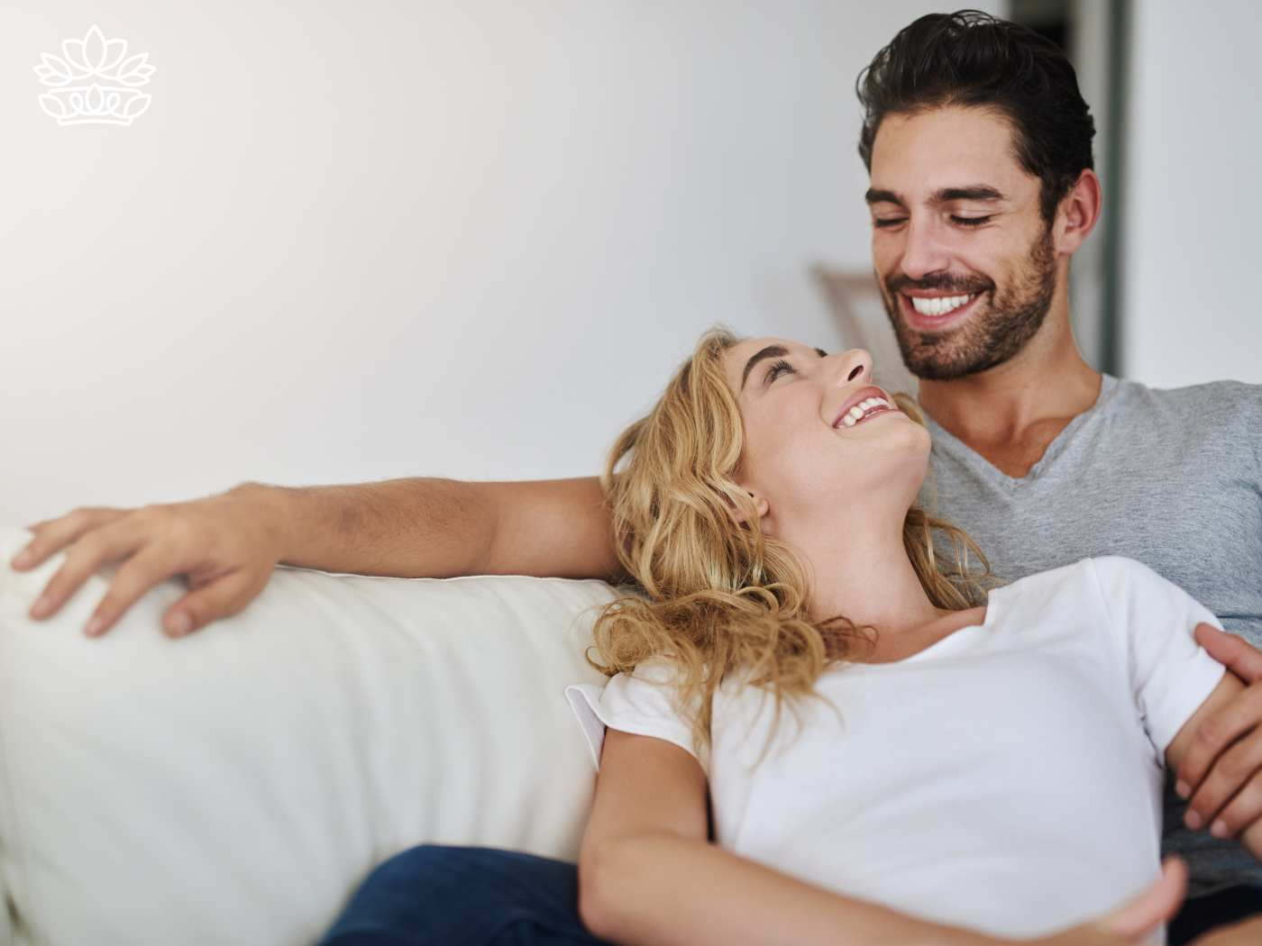 A joyful couple relaxing on a sofa, sharing a laugh in a cozy, bright living room. The man is embracing the woman, who lies back comfortably, reflecting a moment of genuine happiness and intimacy. Perfect scene for celebrating love with Fabulous Flowers and Gifts. Gift Boxes for wife. Delivered with Heart.