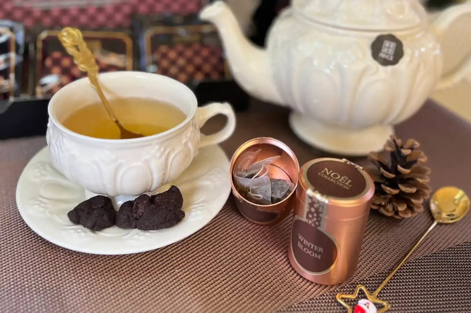 An elegant tea setup on a brown woven mat features a white embossed teacup filled with light tea and two dark cookies on its saucer, paired with a matching large white teapot. Beside the cup, an open rose-gold mini tin reveals pyramid tea bags, positioned next to a pinecone and a decorative gold spoon.