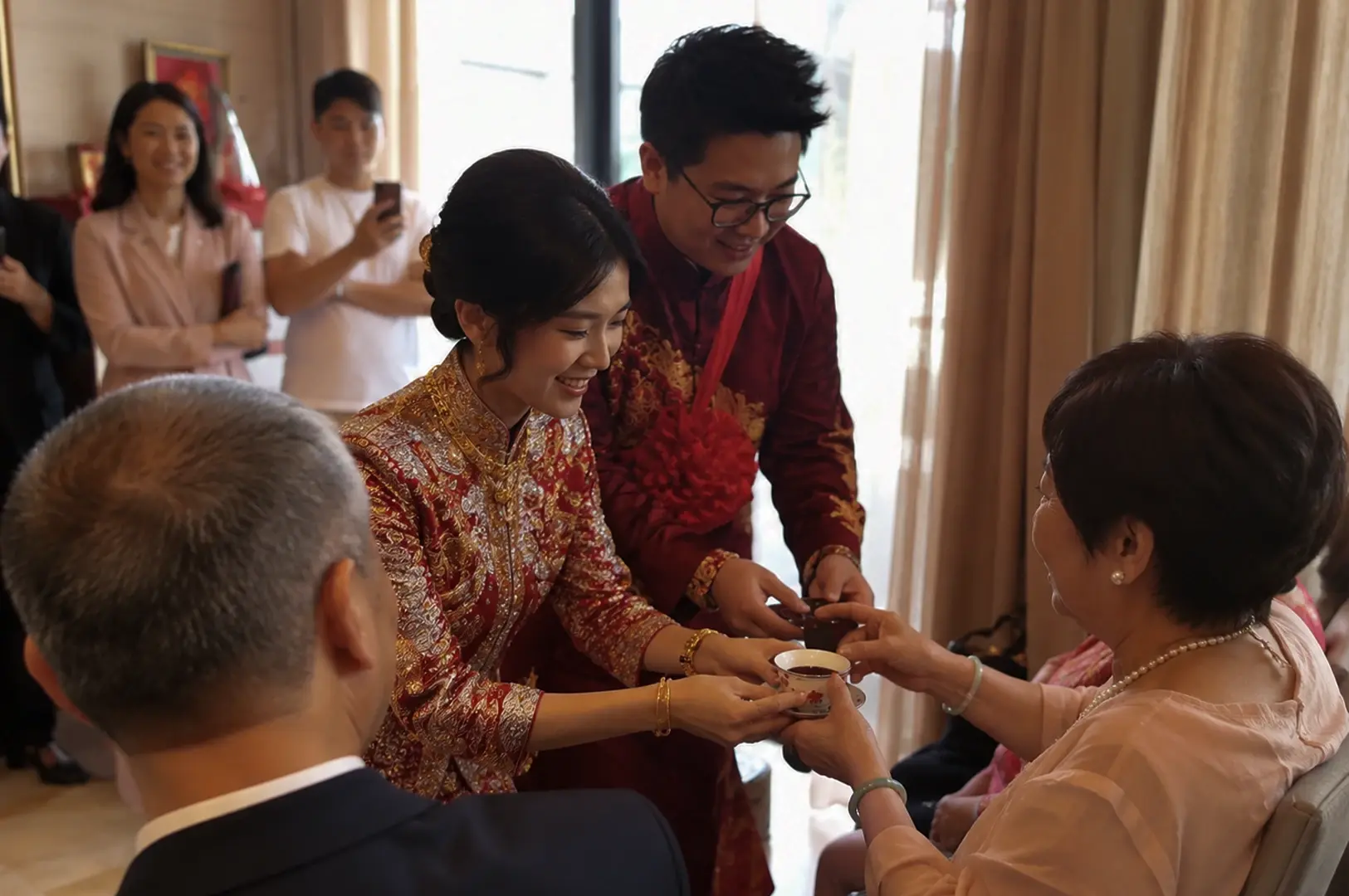 A bride and groom stand close together, smiling warmly as they offer a small white teacup to an older woman seated to the right. The bride wears a red wedding gown covered in dense gold embroidery and accessorized with gold bangles and a gold necklace, while the groom stands beside her in a matching burgundy robe with a large red fabric flower pinned to his chest. The older woman, seen from the side, wears a light pink blouse, a pearl necklace, and jade bangles as she accepts the cup. In the blurred background, other wedding guests look on, including a man in a white t-shirt holding up a phone to take a photograph.