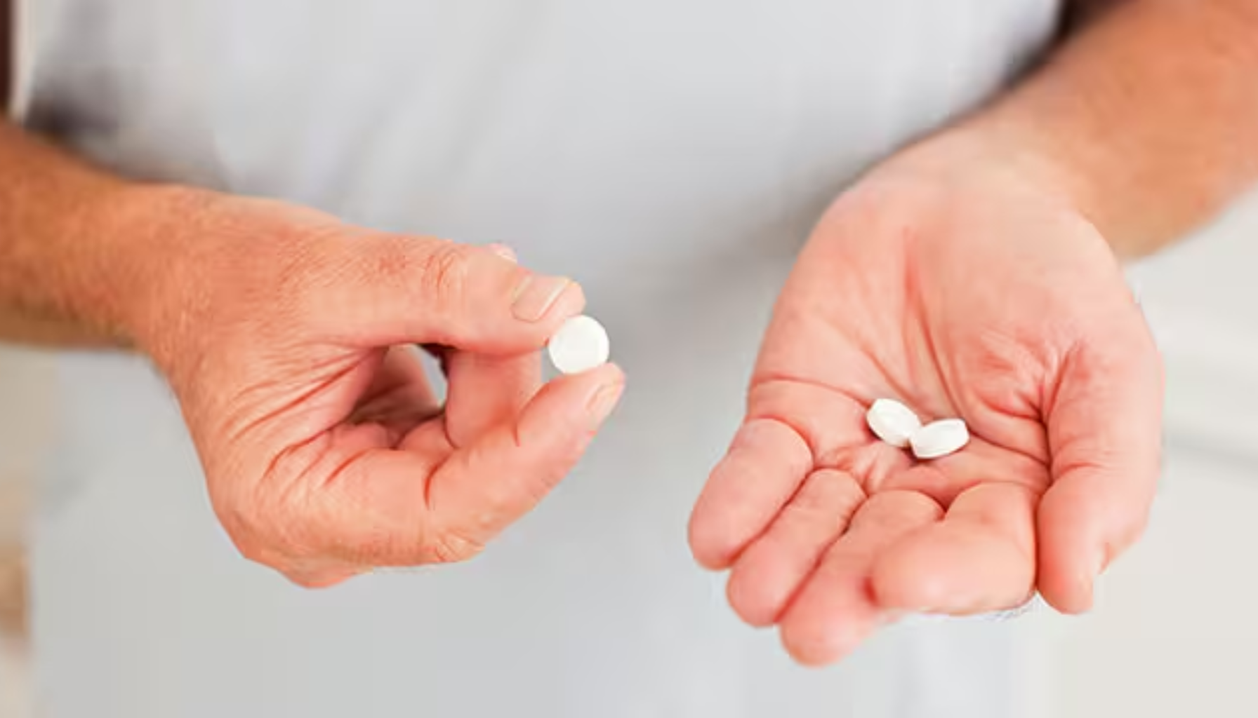 Person holder hvite tabletter i den ene hånden og én pille mellom fingrene foran en uskarp bakgrunn.