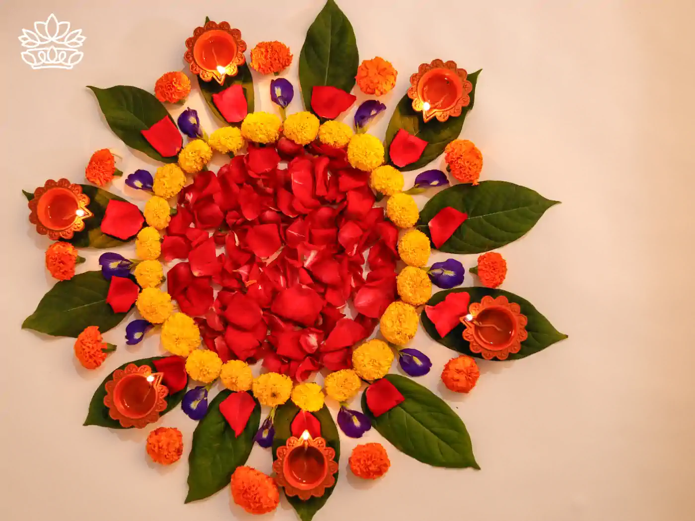 An overhead view of a meticulously arranged Diwali rangoli featuring red rose petals, purple petals, yellow and orange marigolds, interspersed with green leaves and lit clay lamps. Fabulous Flowers and Gifts - Diwali Flowers. Delivered with Heart.