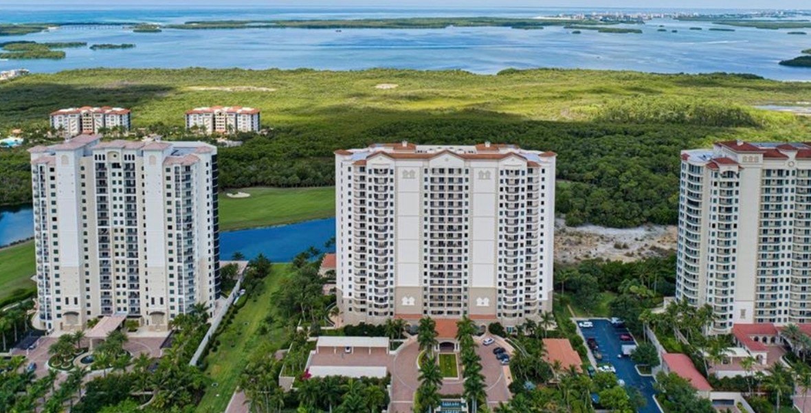 The image showcases a stunning luxury estate home with breathtaking panoramic views of a championship golf course and serene water, featuring an expansive screened lanai and floor-to-ceiling windows that invite natural light into the spacious open floor plan. This premier residence is situated in the exclusive Colony at Pelican Landing, offering access to world-class amenities and a private beach park for residents.