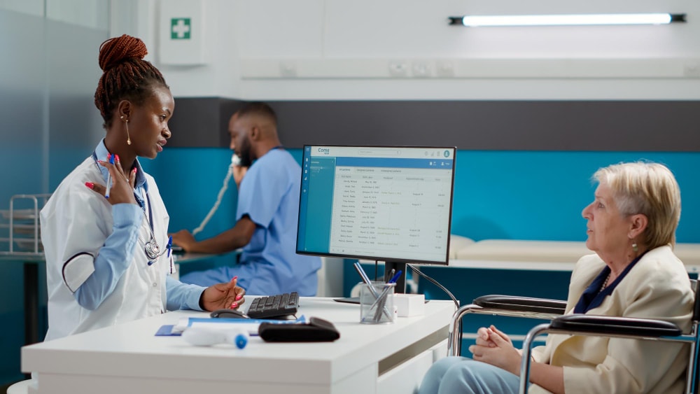 A female doctor at a computer desk speaks with a senior patient in a wheelchair, while another healthcare worker talks on the phone in the background. The use of Unified Communications and team collaboration tools ensures seamless communication between staff and patients, improving business productivity through integrated scheduling and record access.