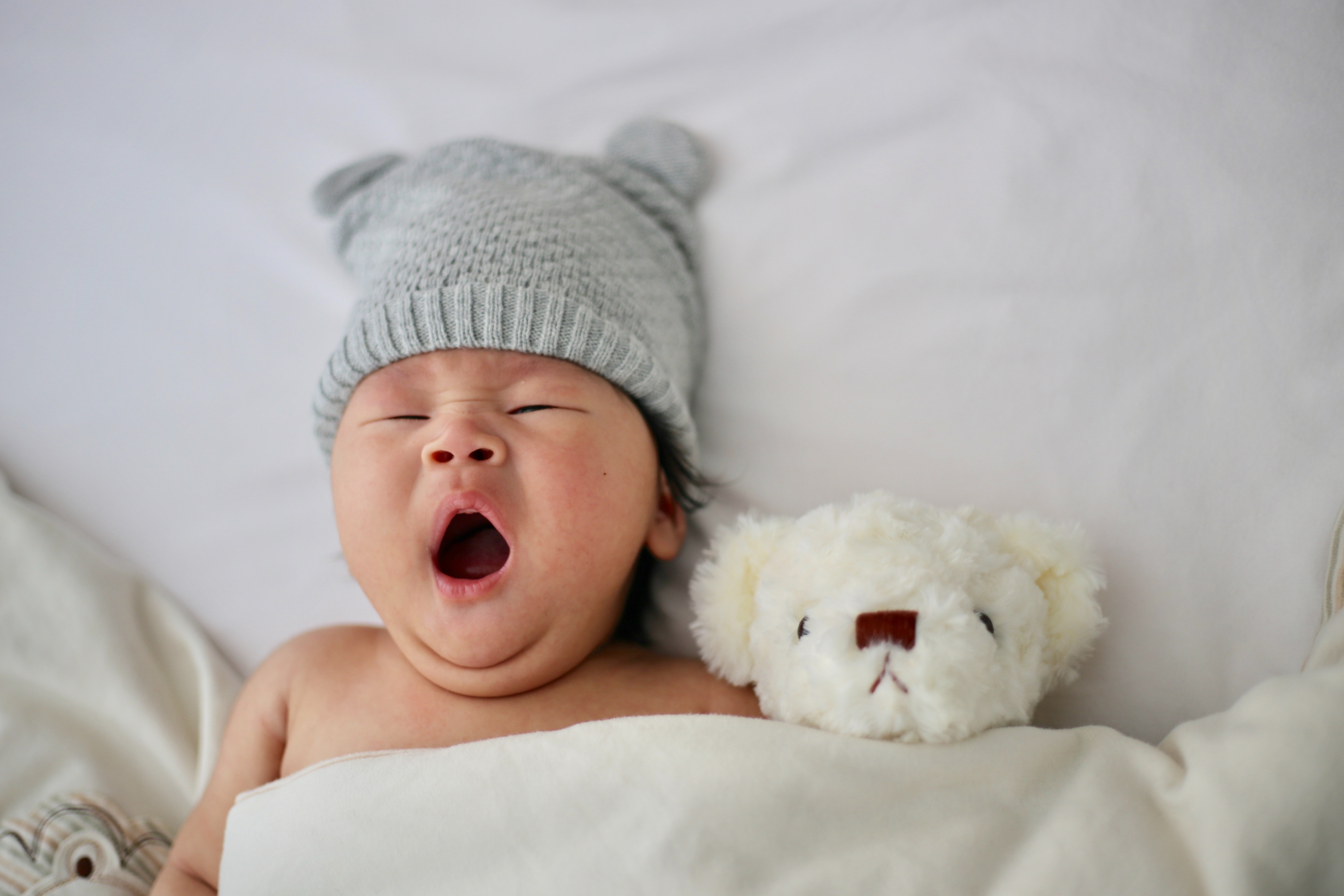 A yawning baby with cute baby's ears laying in a bed tucked in to a blanket, with a teddy bear. 