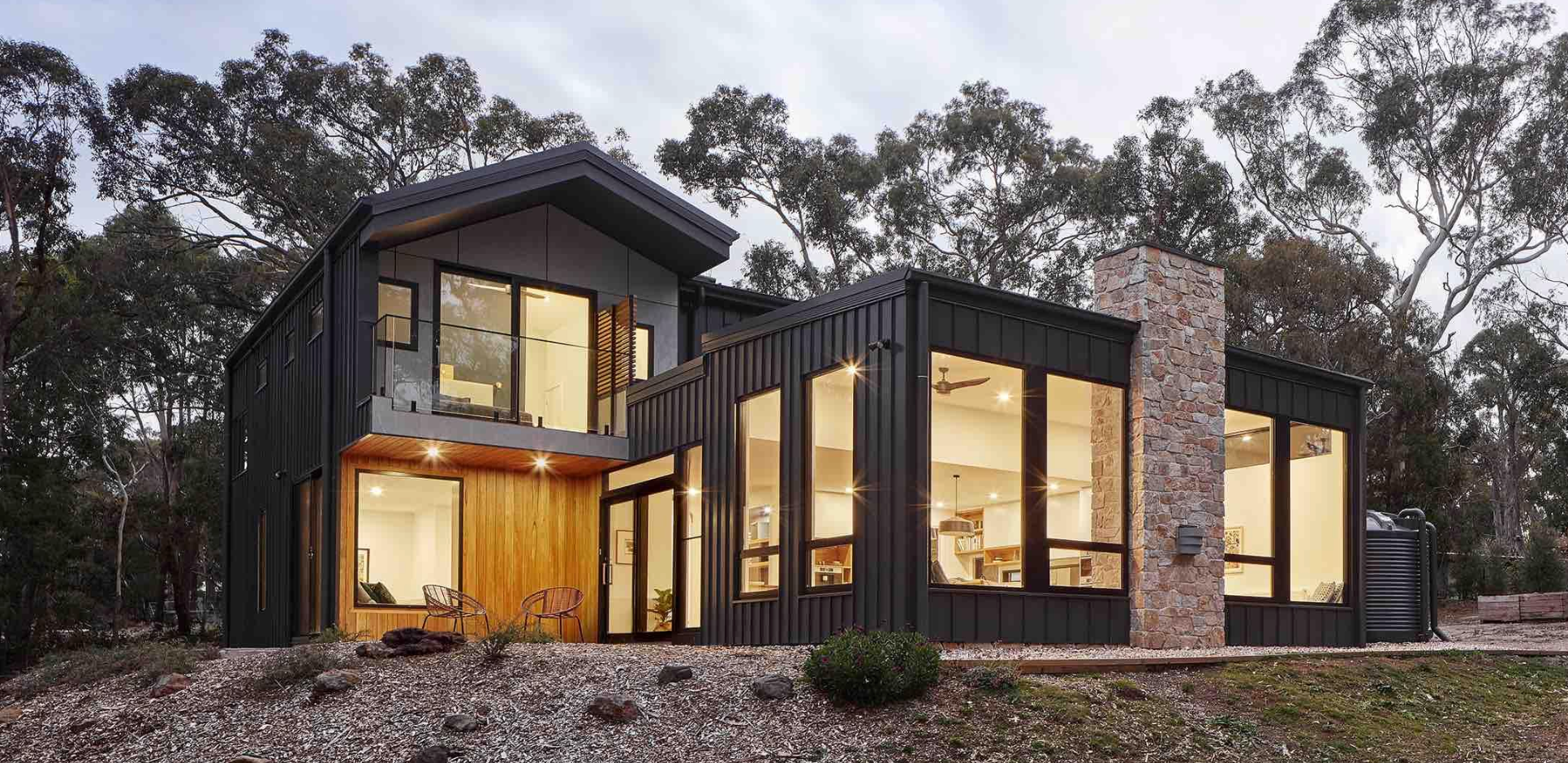 Contemporary home featuring Colorbond steel cladding with crisp and clean lines, large windows, long straight runs of klip lok roofing, a stone chimney, and natural wood accents, set in a forested landscape."