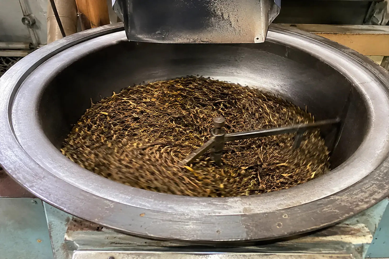 A high-angle close-up shows a large, industrial-style circular metal roaster containing a dark, swirling mass of drying tea leaves, which are being continuously agitated by a rotating mechanical arm.