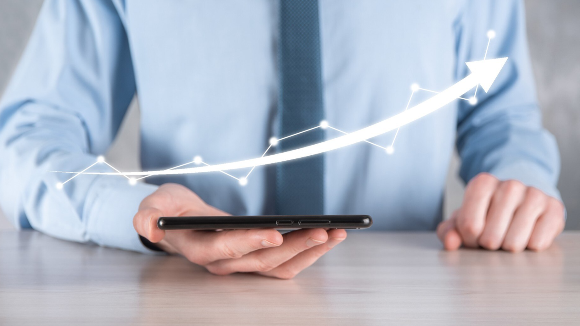 A businessperson in a blue shirt holds a smartphone on a desk, with a glowing upward arrow graph rising from the device. This visual symbolizes business growth powered by mobile communication and Business SMS strategies.