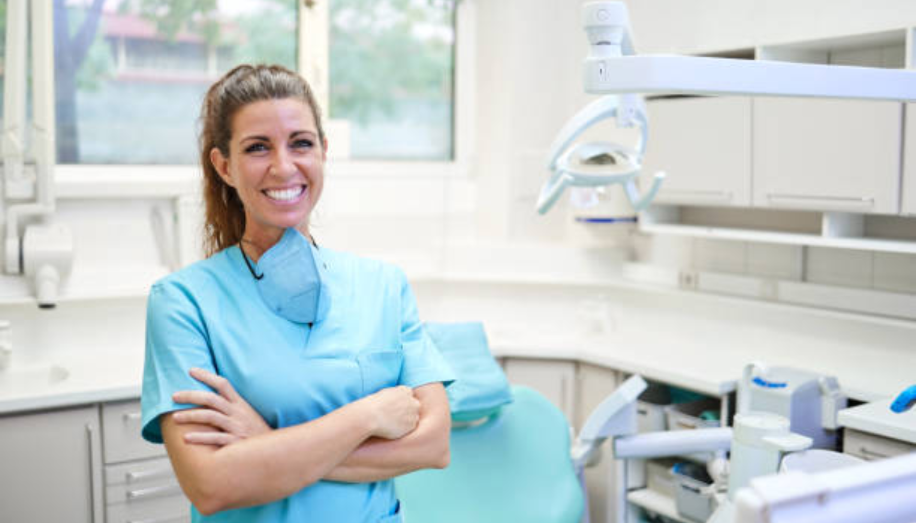 Smilende kvinnelig tannlege i blått uniform står med armene i kors i lyst og moderne tannlegekontor.»
