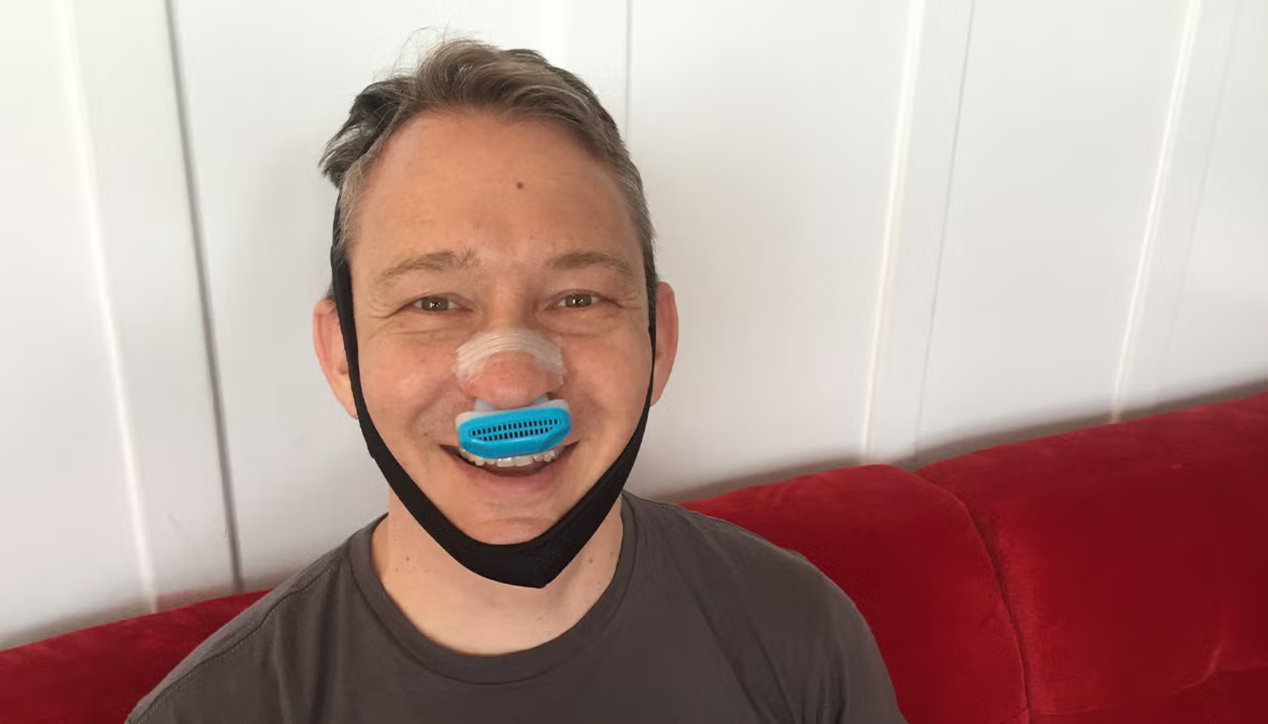 Mann smiler mens han tester blå neseklips og snorkebånd designet for å redusere snorking.