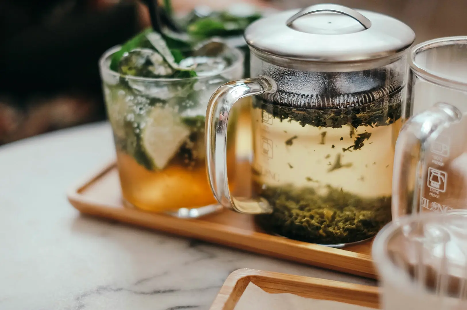 A close-up shot shows a clear glass teapot filled with brewed tea and loose tea leaves sitting on a light wooden tray. Beside it, there is a glass filled with a refreshing iced drink, garnished with fresh mint leaves and citrus slices, all arranged on a light-colored, marbled surface.