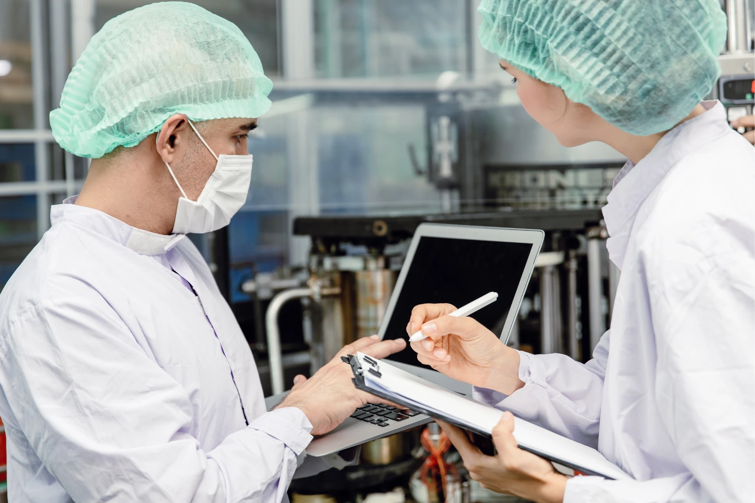 Two people in lab coats and surgical caps reviewing data on a laptop in a manufacturing environment.