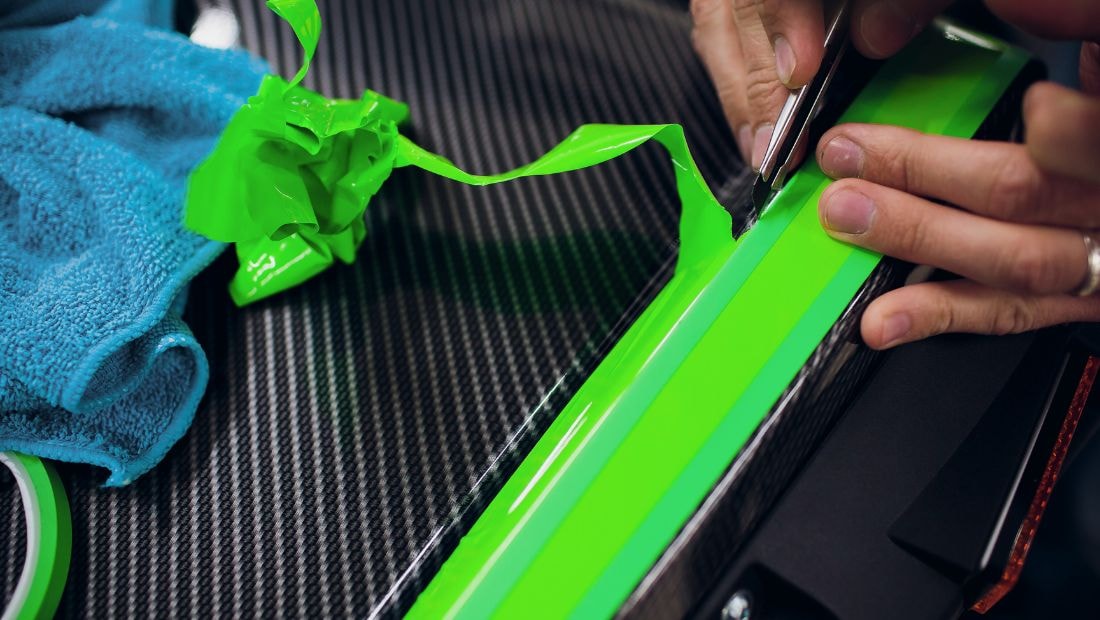 Close-up of hands trimming excess green vinyl wrap with a blade during the detailing phase of a cargo van wrap design, showcasing precision and care.