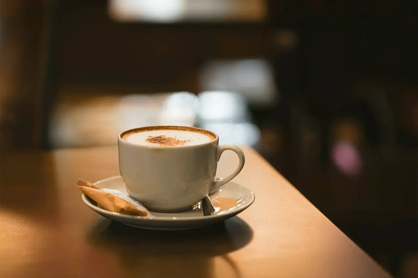 A white coffee cup on a white saucer placed on a smooth wooden table with the dim background blurred behind.
