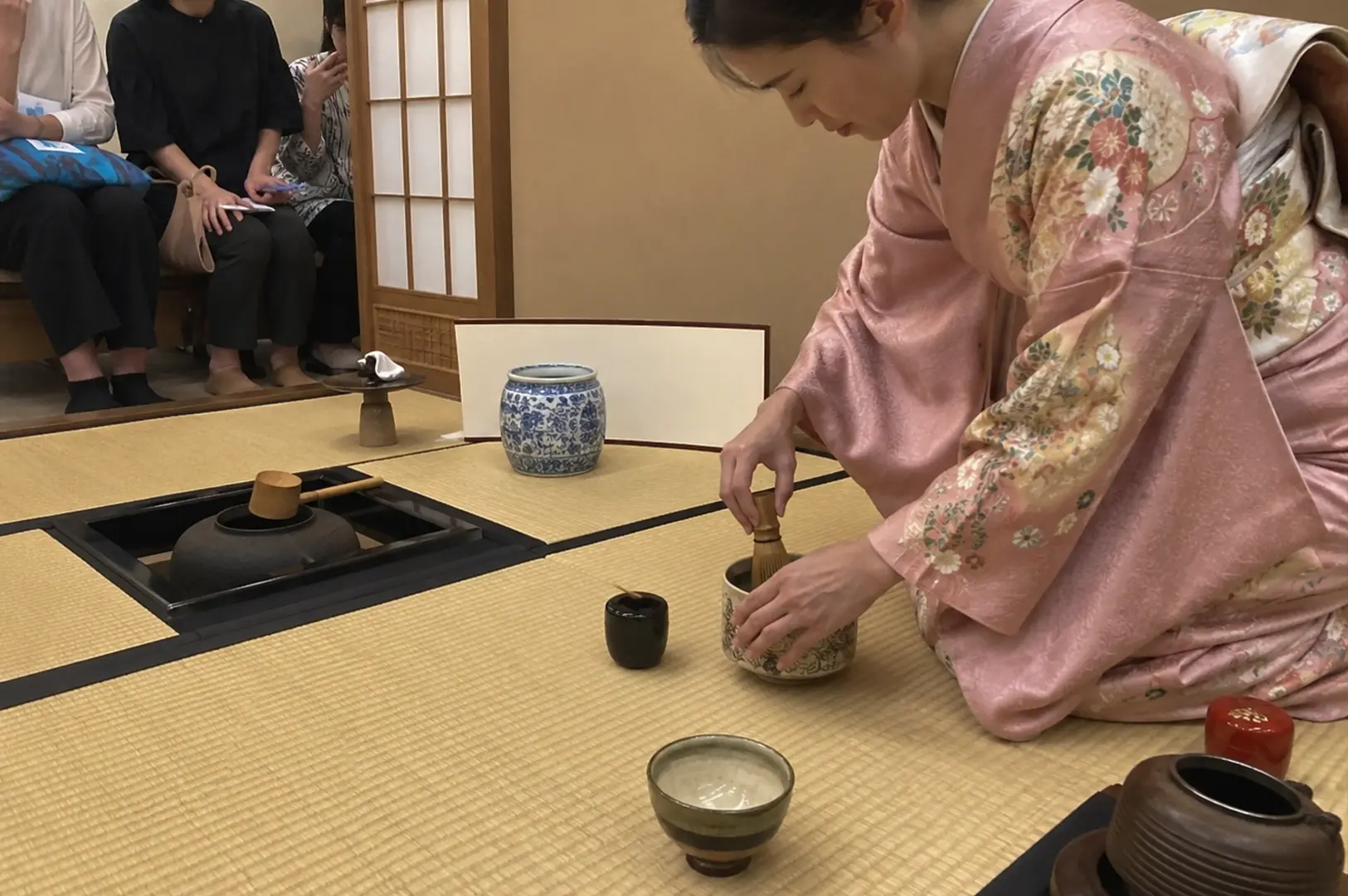 A Japanese tea ceremony unfolds on traditional tatami mat flooring, where a woman dressed in a delicate pink floral kimono kneels gracefully as she whisks matcha inside a large ceramic tea bowl. Various authentic tea utensils are meticulously arranged around her on the mats, including a blue and white patterned ceramic water jar, a black lacquer tea caddy, a wooden water ladle resting over a sunken hearth, and a small dark ceramic bowl in the foreground. In the background, partially separated by a paper shoji screen, a few guests sit on a low bench, quietly observing the practitioner's precise movements.