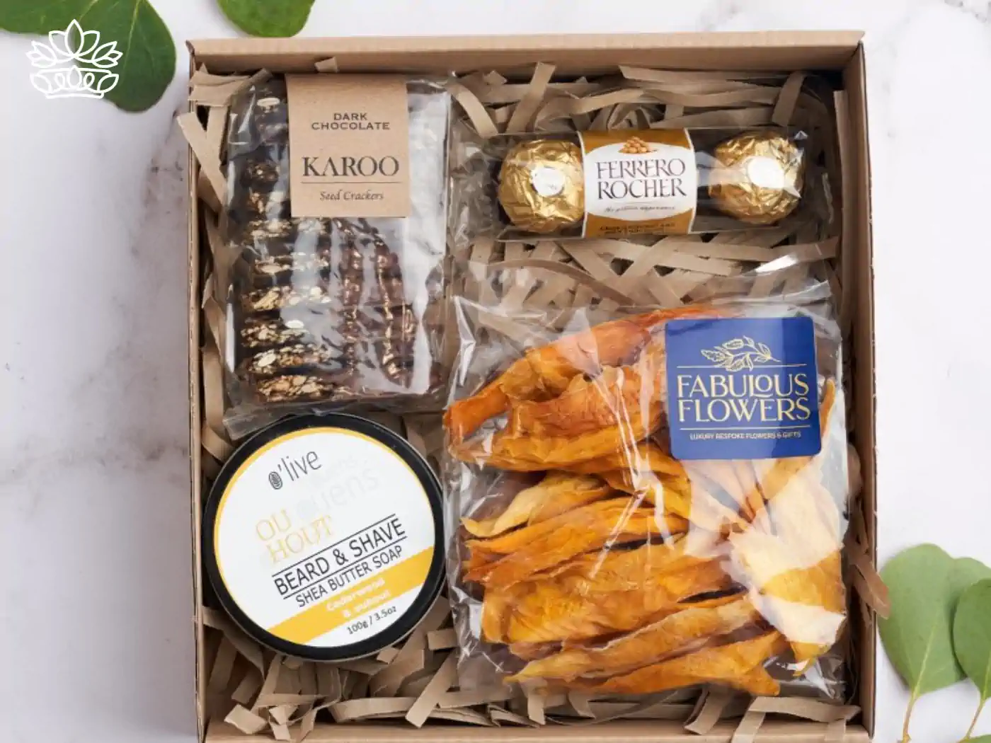 A curated housewarming gift box filled with luxurious items, including dark chocolate seed crackers, two Ferrero Rocher chocolates, a container of beard and shave butter soap, and dried mango slices, all arranged in a rustic box with straw padding. Thoughtfully selected for a warm welcome home. Fabulous Flowers and Gifts.