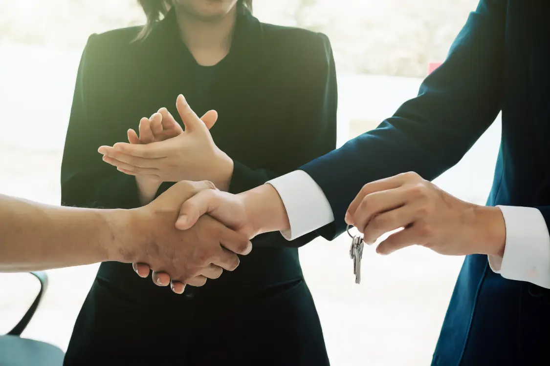 Businesspeople shaking hands while handing over keys during a deal closing, the final step after a successful interview using key interview questions for business brokers.